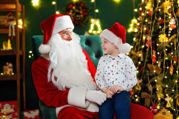 Laughing little boy in Santa's hat sitting with Santa Claus near Christmas tree. New Year concept 
