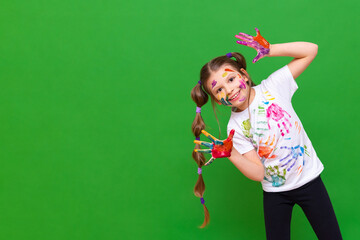 A little joyful girl with stained hands in paint makes faces and smiles, a little girl got dirty in a rack on a green isolated background.