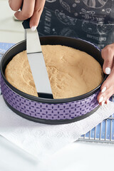Cropped close-up shot of woman's hands with a rounded knife removing a pie from a cake tin on a table in the kitchen. Cake pan has purple fabric strips. Top view.