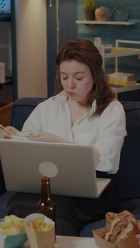 Vertical video: Young woman using laptop on sofa while eating asian food with chopsticks. Caucasian person sitting at home with chinese takeaway meal, looking at movie on TV and working on gadget