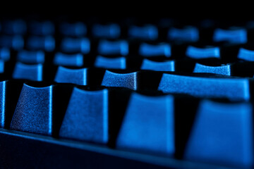 Gaming computer keyboard with black keycaps illuminated in the dark by blue light from a monitor or...