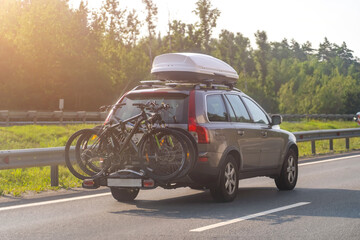 Bikes fastened on bicycle holder mounted on back side of car on country road. Brown car with roof...