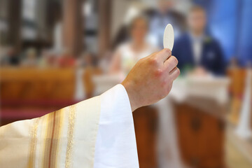 Holy host in the hands of the priest on the altar during the celebration of the wedding mass on the background of the bride and groom