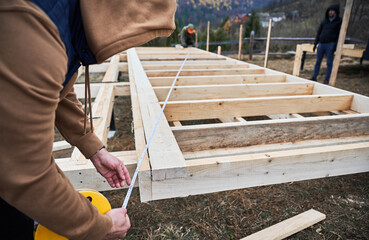 Man worker building wooden frame house on pile foundation. Carpenter using tape measure for...