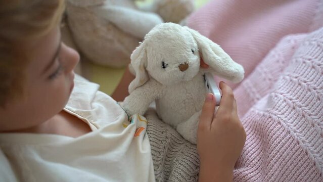 Sick child playing with his ill toy hare, measure body temperature with electronic thermometer. Role playing, child playing doctor with plush toy. Children cold and flu, illness concept. Top view