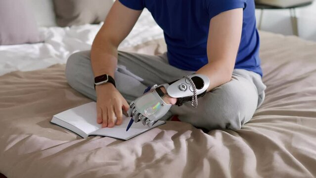 Close up shot of man with prosthetic bionic hand writing with pen in notepad sitting on bed