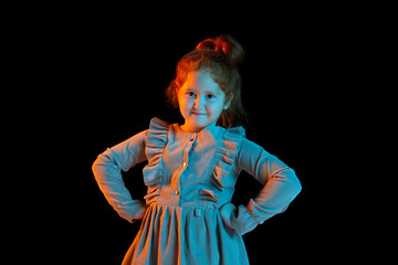 Half-length portrait of cute redheaded little girl wearing festive dress isolated over dark background in neon light. Kids emotions, facial expressions, fashion