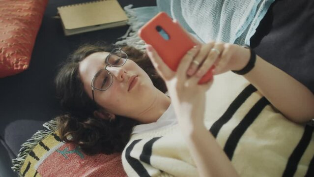 High angle shot of girl lying on sofa at home and surfing the Internet on mobile phone