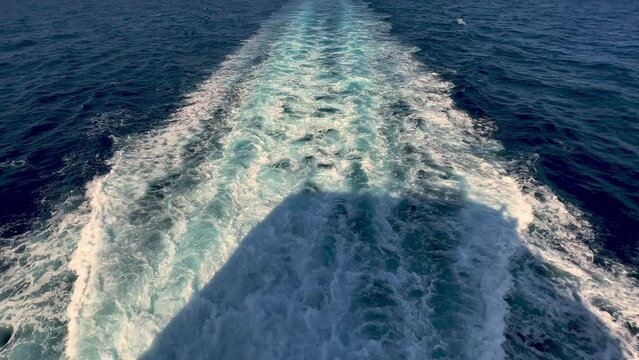 Rear view of wide water wake left from cruise ship on sea water surface with horizon in background and shadow of people leaning over parapet of deck