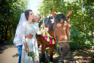 Fototapeta premium Stylish groom in a blue suit walk with the bride in a white dress in nature in the park with white horse. Wedding portrait of happy newlyweds on a background of greenery