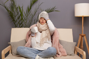 Portrait of unhealthy sick Caucasian woman wearing warm clothes and wrapped in scarf sitting with cup of hot tea in hands, feels bad, having high temperature, suffering terrible headache.