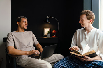 Happy multiracial gay couple using laptop and reading book at home