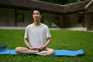  Portrait of Millennial man practicing and sitting in lotus asana pose on the grass field. © saltdium