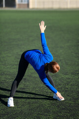 Women and sport. Girl in sportswear - blue shirt and black leggings does exercises: bends and stretches on the grass at the stadium on a sunny day. Middle aged sportswoman 
