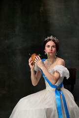 Fast food or dieting. Young funny girl like queen or princess in white medieval outfit eating burger on dark background. Concept of comparison of eras, modern, fashion, beauty. © master1305