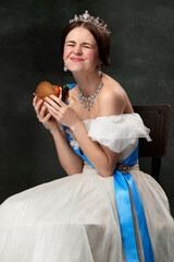 Emotional young woman, royal person, queen or princess in white medieval outfit eating burger on dark background. Concept of comparison of eras, modern, fashion, beauty.