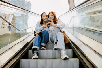 Young multinational women using cellphone while sitting on escalator