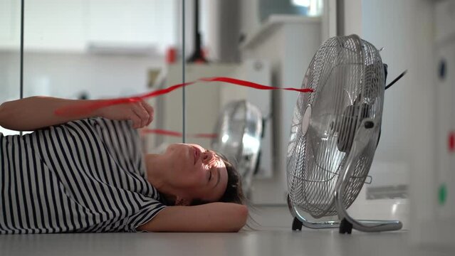Cheerful young Caucasian woman lies on floor in front of working electric fan with red ribbon. Lonely girl rejoices in cold wind being in apartment during abnormally hot and sunny weather outside