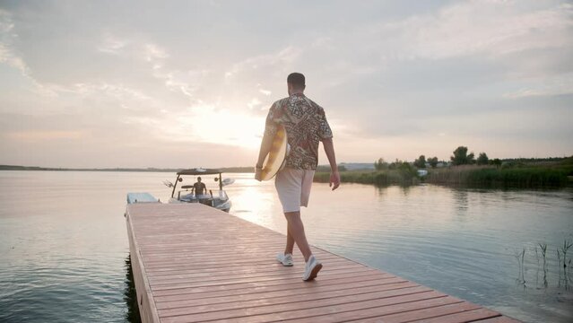 Rear of young casual stylish man walking on pier on lake on sunset carrying water board in hand. Male surfer walks on jetty to white motor boat with small surf board in hands. Summer water sport