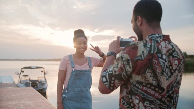 Close up of happy young woman smiling while posing to camera outdoor near lake. African American boyfriend taking photos of his girlfriend on retro photo camera standing in nature on lake. Photography