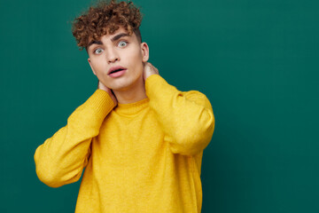 Obraz na płótnie Canvas a shocked man in a yellow sweater stands on a green background and holds his head with his hand, his mouth wide open, looking into the camera from shock