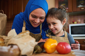 Loving Middle Easter Muslim mother, beautiful smiling woman in hijab and her cute little daughter, standing close to each other while unpacking groceries from a shopping bag in the kitchen at home