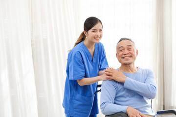 The caregiver therapist stands with an Asian senior sitting in a wheelchair and touches their hands on senior shoulder together. The nursing home facilitates a support group. Image with copy space