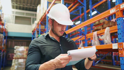 Close-up of Caucasian logistics employee looking at paperwork to check the amount of cargo cardboard boxes on shelves in large warehouse. Logistics manager concentrate on work with the stock of goods.