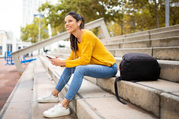 happy young woman sitting with mobile phone listening to music with earbuds