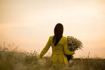 Fototapeta premium a girl in a yellow dress at sunset in a chamomile field with a bouquet of daisies
