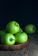 Green apples are placed in a wooden tray and rest on a rustic wooden floor.