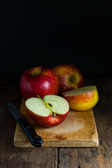 Red apples, whole, and halves with a kitchen knife put on a wooden cutting board on a rustic wooden floor.