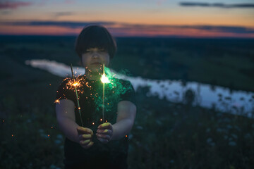 A cute boy holds sparklers in his hands, fireworks in the evening in the summer at sunset. Festive mood.