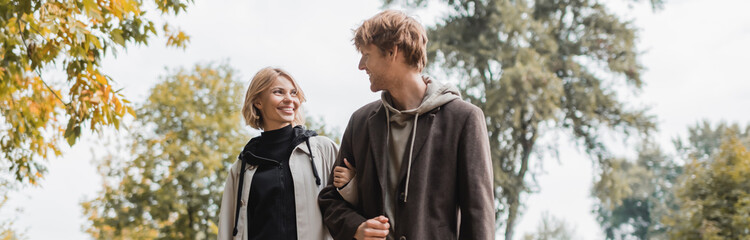 positive couple in coats looking at each other while walking in autumnal park during date, banner.