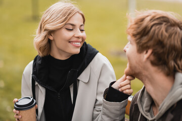 cheerful blonde woman holding paper cup and touching chin of redhead boyfriend.