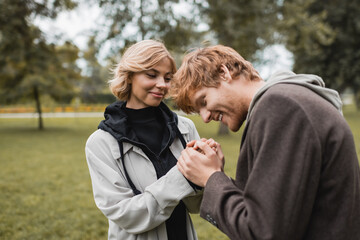 caring young man in coat warming up hands of happy girlfriend in autumnal park.