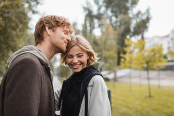 happy redhead man kissing head of blonde girlfriend smiling in park.