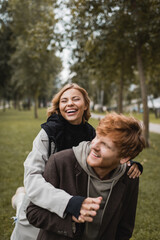 positive young woman in coat hugging cheerful redhead boyfriend and laughing in autumnal park.