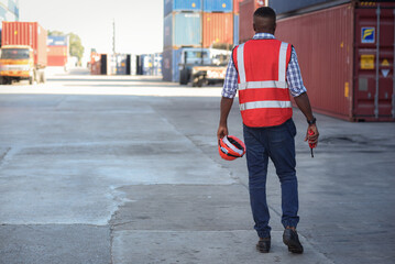 Portrait back side of an African-American male worker in safety uniform workwear Hold a walkie-talkie and hardhat.Walking into  warehouse to stock inspection, containers lined up in goods loading area