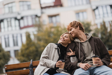 smiling young woman hugging man and holding coffee to go while sitting on bench in park.