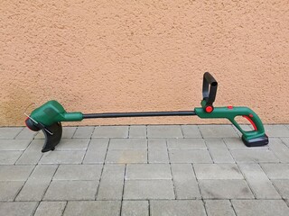 String electric lawnmower, trimmer, green plastic body, placed on concrete pavement by the wall of the house