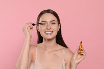 Young woman applying oil onto her eyelashes on pink background