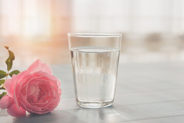 A glass of clear drinking water with a good morning message and pink rose in the morning with smooth sunlight, Concept Drinking clean water in the morning for good health, Good start in the morning.
