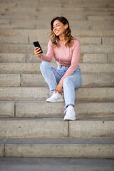 Full body happy woman sitting on stairs with cellphone and earphones