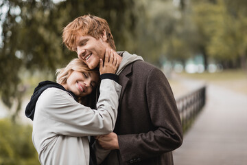 pleased redhead man and blonde woman in coat smiling while having date in park.