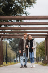 full length of joyful couple in autumnal coats holding hands while walking under multiple arch in park.