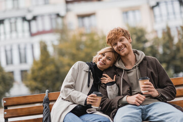 cheerful young woman hugging man and holding coffee to go while sitting on bench in park.