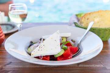 Traditional Greek Salad served in tavern, traditional greece food with Aegean sea as background. Tomatoes, cucumber, onions, olives, peppers, cappers and olive oil