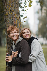 happy blonde woman holding paper cup and hugging with boyfriend near tree trunk.
