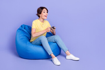 Full body portrait of overjoyed positive lady sitting comfy bag play games isolated on purple color background